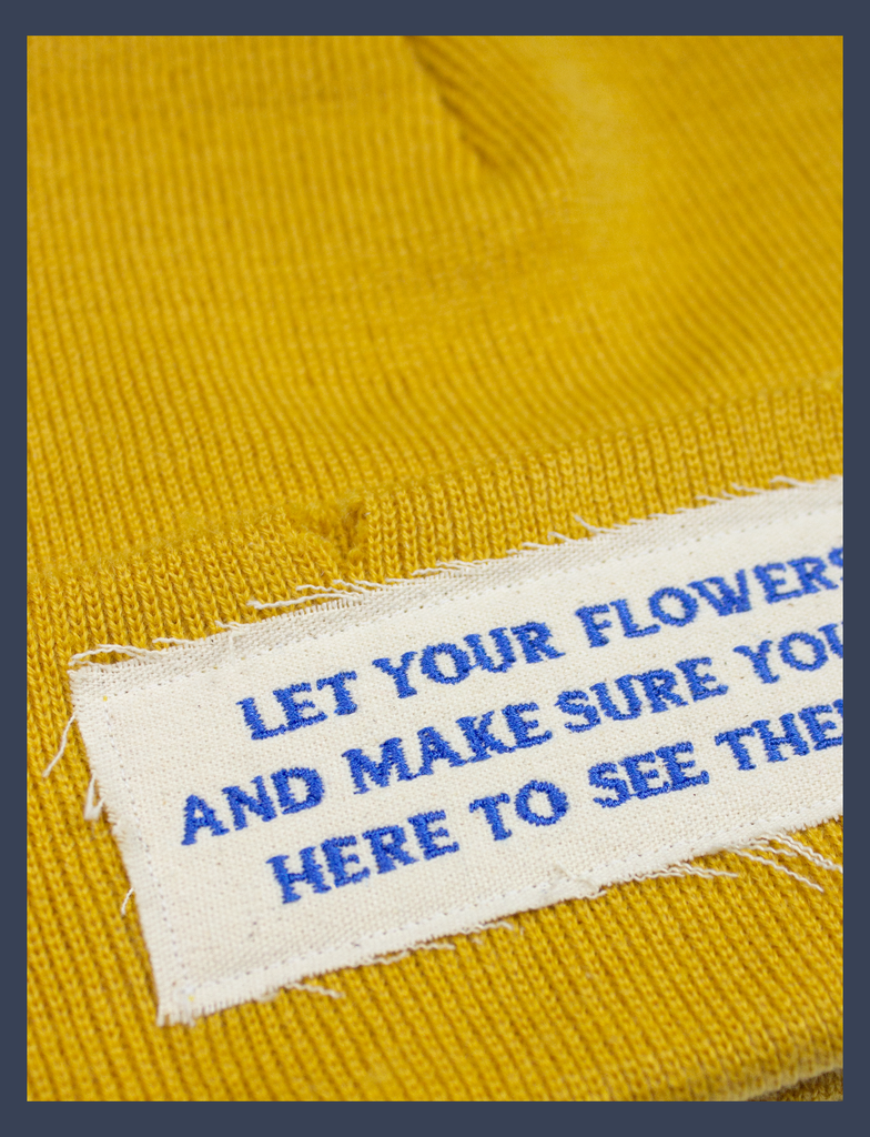 Close-up of the embroidered lettering on the FLOWERS Beanie in Mustard Yellow by KULT Clothing | The speckled appliqué label reads, "Let your flowers grow and make sure you are still here to see them bloom." | 100% acrylic knit beanie hat with distressing across it's construction and design