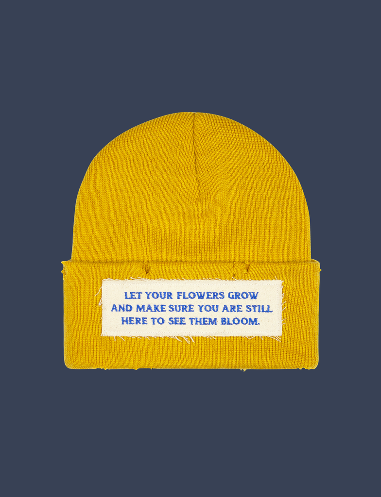 Front of the FLOWERS Beanie in Mustard Yellow by KULT Clothing | It features a speckled cream appliqué label with an embroidered design in cobalt blue thread on the front | The design reads, "Let your flowers grow and make sure you are still here to see them bloom." | 100% acrylic knit beanie hat with distressing across it's construction and design