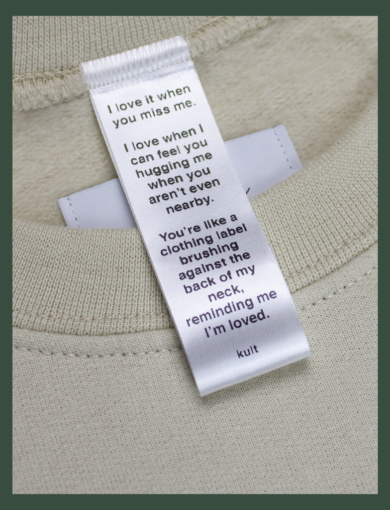 Close-up on the label of the DISTANT LOVE Sweater in Sandstone by KULT Clothing | I love it when you miss me. I love when I can feel you hugging me when you aren’t even nearby. You’re like a clothing label brushing against the back of my neck, reminding me I’m loved. KULT