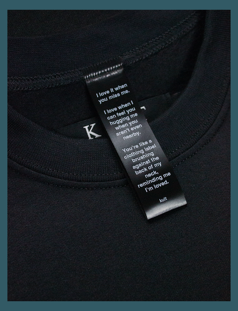 Close-up on the label of the DISTANT LOVE Sweater in Black by KULT Clothing | I love it when you miss me. I love when I can feel you hugging me when you aren’t even nearby. You’re like a clothing label brushing against the back of my neck, reminding me I’m loved. KULT