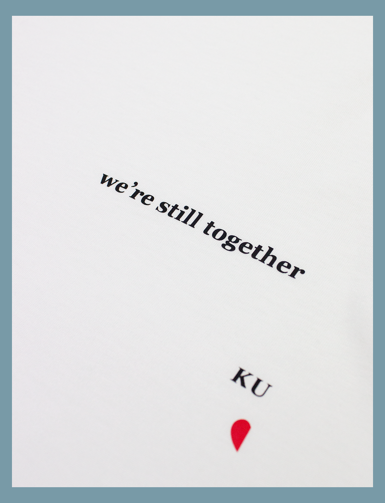 Close-up view showing part of the design on the ALWAYS TOGETHER Tee in White by KULT Clothing | It reads "We're still together" and shows half of the KULT logo and heart symbol printed across the left breast using our matte black and red Barely There™ vinyl | eco-friendly, climate neutral t-shirt