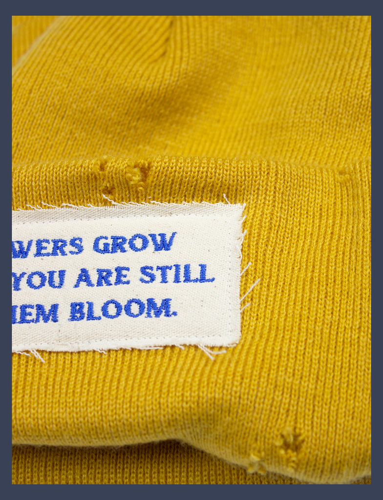 Close-up on the hand-distressed detailing across the body of the FLOWERS Beanie in Mustard Yellow by KULT Clothing | The speckled appliqué label reads, "Let your flowers grow and make sure you are still here to see them bloom." made using embroidered lettering | 100% acrylic knit beanie hat with distressing across it's construction and design