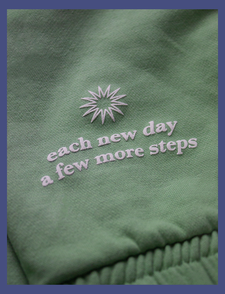 Close-up view of the hand-printed puff design on the EMBRACE Sweatpants in Sage by KULT Clothing | It reads "Each new day / A few more steps" next to a stylised illustration of a sunrise | Hand-printed using eco friendly inks